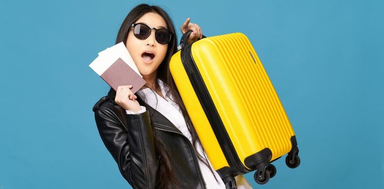 A woman wearing sunglasses and holding a yellow suitcase in one hand, and her passport and plane tickets in the other hand.