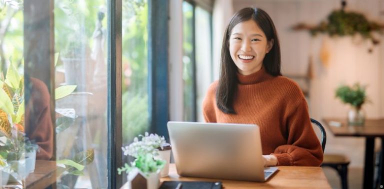 A woman sitting at a table with a laptop. She's wearing an orange sweater and smiling.