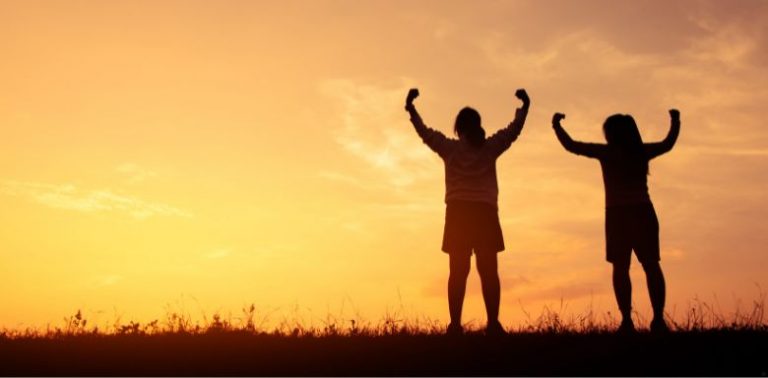 Two people standing outside together with their arms up in the air, in front of a sunset.